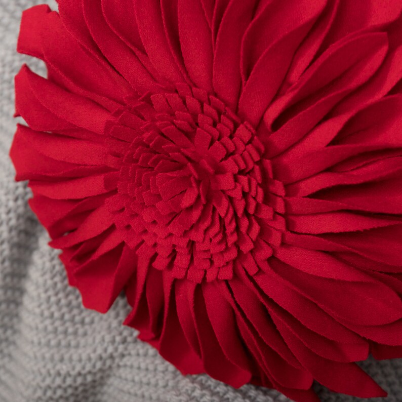 May include: A red felt flower pillow with a large, multi-layered center. The flower is in focus and the background is a blurry gray.