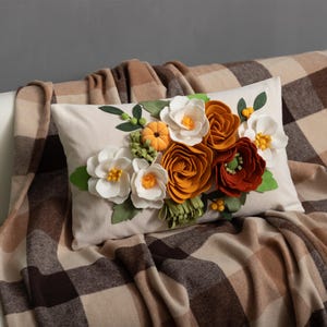 Puede incluir: Un cojín decorativo rectangular con un diseño floral. El cojín presenta flores de fieltro blancas, naranjas y rojas, hojas verdes y pequeñas calabazas naranjas. El cojín está sobre una manta a cuadros.