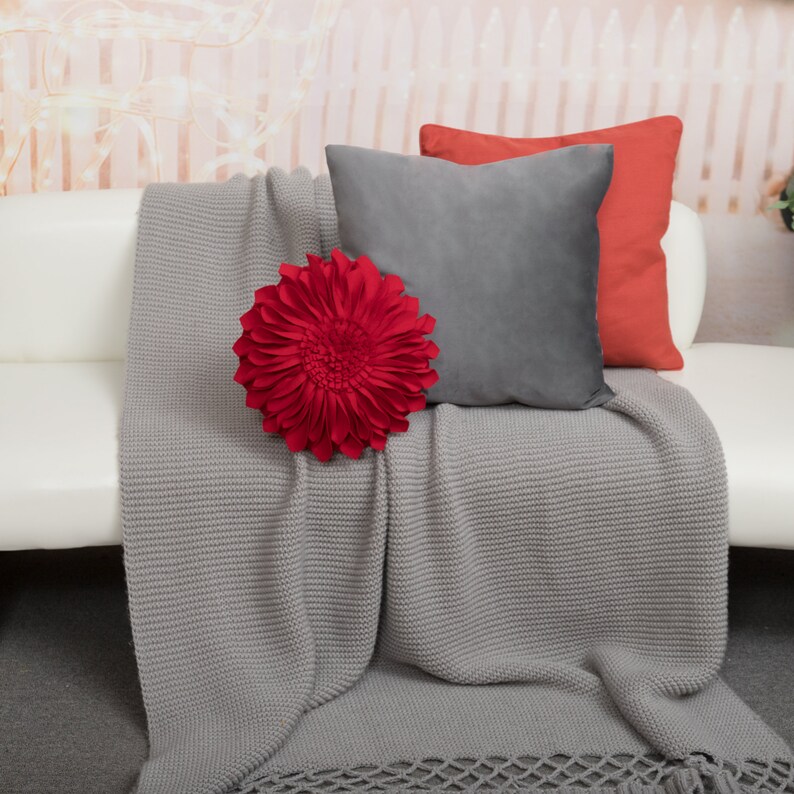 May include: A gray knitted throw blanket with a red flower-shaped pillow on top. Two gray and red pillows are behind the flower pillow.