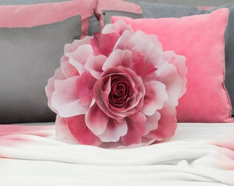 May include: A large pink flower pillow with a white center, sitting on a bed with pink and gray pillows.