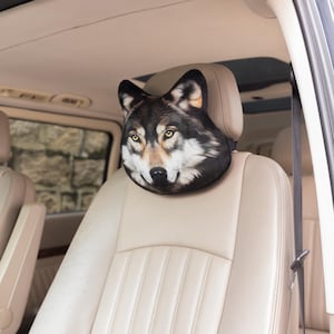 May include: A car headrest cover shaped like a wolf's head. The headrest cover features a realistic depiction of a wolf with black, gray, and white fur, and yellow eyes. The headrest is placed on a beige car seat.