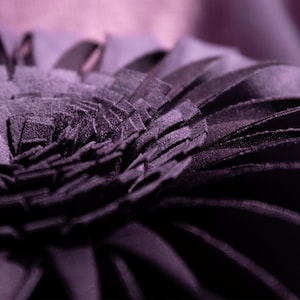 May include: A deep purple decorative pillow featuring a layered, petal-like design constructed from numerous fabric strips. The circular arrangement creates a dense, textured surface that radiates from the center, showcasing a dark violet hue.