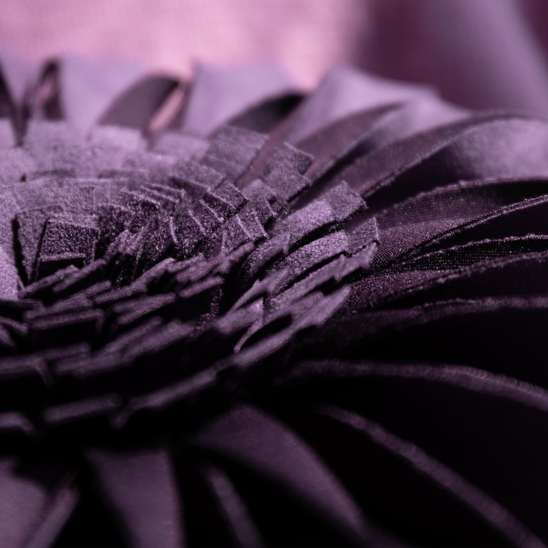 May include: A deep purple decorative pillow featuring a layered, petal-like design constructed from numerous fabric strips. The circular arrangement creates a dense, textured surface that radiates from the center, showcasing a dark violet hue.