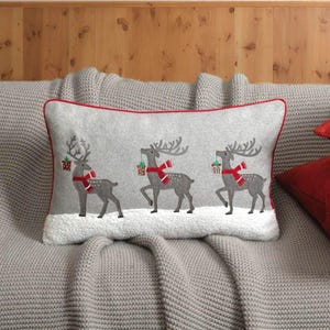 May include: Rectangular gray decorative pillow featuring three reindeer with red scarves and holding gifts. The pillow has a red border and is placed on a gray knitted blanket. The reindeer are gray with embroidered details.