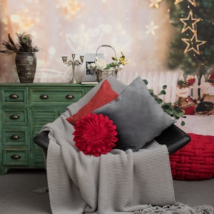 May include: A gray knitted throw blanket draped over a black armchair with three pillows. One pillow is red with a large flower design, one is gray, and one is red. A red braided ottoman sits in the foreground.