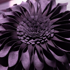 May include: A deep purple decorative pillow shaped like a blooming flower. The pillow features numerous layered, petal-like fabric strips radiating from a dense, textured center composed of many small, upright rectangular fabric pieces.