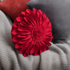 May include: Red felt flower pillow with a large, round, three-dimensional flower design. The pillow is on a gray knitted throw blanket.