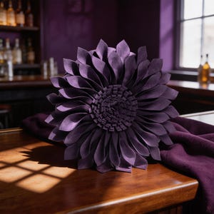 May include: A round, deep purple decorative pillow shaped like a blooming flower. The pillow features multiple layers of pointed, petal-like fabric strips radiating from a dense, fringed center, creating a textured, three-dimensional floral appearance.