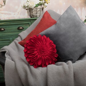 May include: A red flower-shaped throw pillow with a gray and red throw blanket and two decorative pillows on a couch.