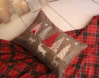 May include: A decorative Christmas pillow with a brown background and a red border. The pillow features embroidered Christmas trees, reindeer, and snowflakes. It rests on a red and black plaid blanket, creating a festive holiday scene.