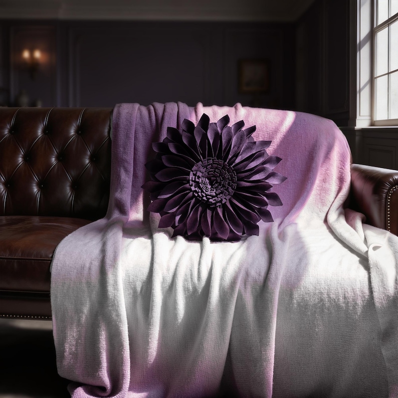 May include: A dark purple, flower-shaped throw pillow with layered, pointed petals sits atop a soft, gradient throw blanket that transitions from a light lavender hue at the top to a crisp white color at the bottom, draped over a dark brown sofa.