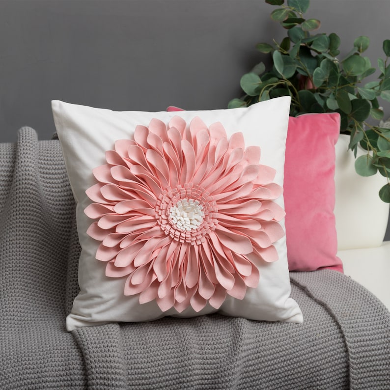 May include: A white pillow with a large pink flower design. The flower is made of fabric petals and has a white center. The pillow is sitting on a gray knitted throw blanket.