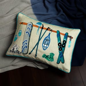 May include: Rectangular beige pillow with embroidered winter sports equipment, including skis, poles, snowshoes, and mittens. The pillow has a teal border and is set against a backdrop of blue and gray blankets.