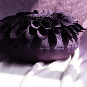 May include: A round, deep purple throw pillow featuring a textured surface with layered, petal-like fabric folds arranged in a circular, flower-inspired pattern across the top. The material has a soft, suede-like finish throughout the entire item.