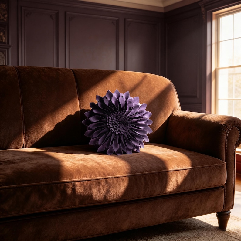 May include: A round, purple, flower-shaped decorative pillow with layered, petal-like fabric folds sits centered on a brown, velvet-textured sofa. The sofa features a smooth, upholstered surface with piped edges and dark, rounded wooden legs.