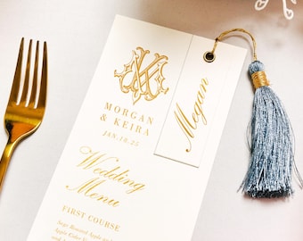 Könnte beinhalten: Eine weiße Hochzeitsmenükarte mit goldener Folienprägung und einem Monogramm mit den Namen Morgan & Keira, datiert auf den 18. Jan. 25. Ein kleineres Schild mit dem Namen Megan ist mit einer goldenen Kordel an einer staubblauen Quaste befestigt. Links liegt eine goldene Gabel.