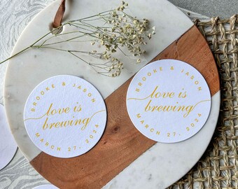 May include: Two white round paper coasters with gold lettering. The coasters say "Brooke & Jason love is brewing" and include the date "March 27, 2024".