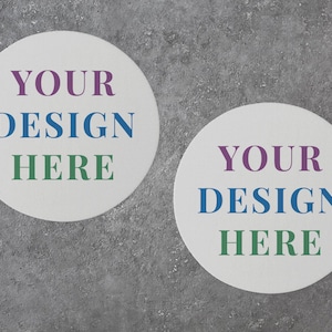 Może przedstawiać: Dwa białe okrągłe podkładki pod kubki z tekstem "YOUR DESIGN HERE" w kolorze fioletowym, niebieskim i zielonym.