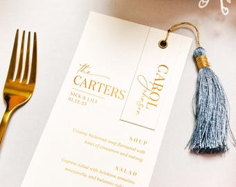 Könnte beinhalten: Eine goldfarbene Gabel liegt neben einer cremefarbenen Hochzeitsmenükarte. Die Karte zeigt goldfarbene Schriftzüge mit den Namen "The Carters" und "Nick & Lily" sowie dem Datum "01.12.25". Ein kleineres Etikett mit dem Namen "Carol Johnson" hängt an der Karte und ist mit einer blauen Quaste verziert.