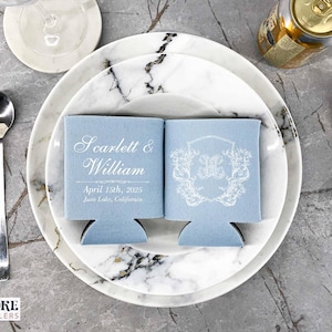 May include: Two light blue can coolers with white text. The left cooler says "Scarlett & William" and "April 15th, 2025" and "June Lake, California". The right cooler has a white floral design with a monogram.