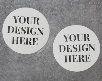 May include: Two white round coasters with black text that says "YOUR DESIGN HERE" on a gray textured surface.