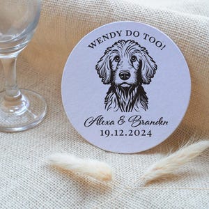 Op de afbeelding: Ronde witte onderzetter met zwarte tekst die "Wendy Do Too!" luidt en een zwart-witte illustratie van een hond. De tekst onder de hond luidt "Alexa & Branden 19.12.2024".