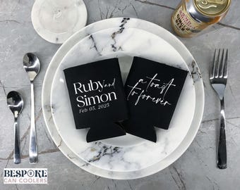 Peut inclure: Deux manchons noirs pour canettes avec texte blanc, l'un indiquant "Ruby and Simon Feb 05, 2025" et l'autre "a toast to forever". Les manchons sont sur une assiette à motif marbré, avec des couverts et une canette de ginger beer.
