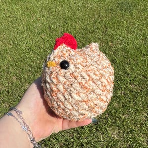 May include: A handmade, round crochet chicken with a red comb, black eye, and yellow beak. The chicken is made of cream and orange textured yarn. The background is green grass.