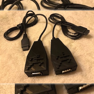 Puede incluir: Dos cables negros con conectores negros y un adaptador negro con un gráfico de extraterrestre pixelado a cada lado.