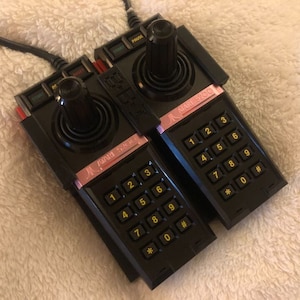 May include: Two black Atari 5200 controllers with pink labels and yellow buttons. Each controller has a joystick and a keypad with numbers and symbols.