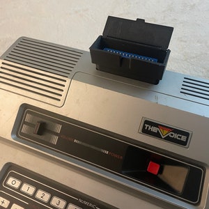 May include: A silver and black The Voice talking computer with a red power button and a black cartridge slot on the top. The cartridge slot has a black lid that is open.