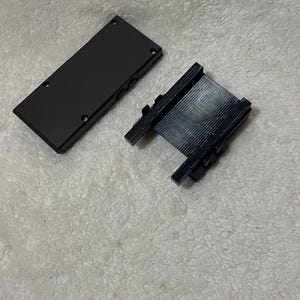 May include: Two black rectangular components, possibly electronic device parts, are displayed against a white, textured background. One component is a solid rectangle with screws, and the other is a frame-like structure.