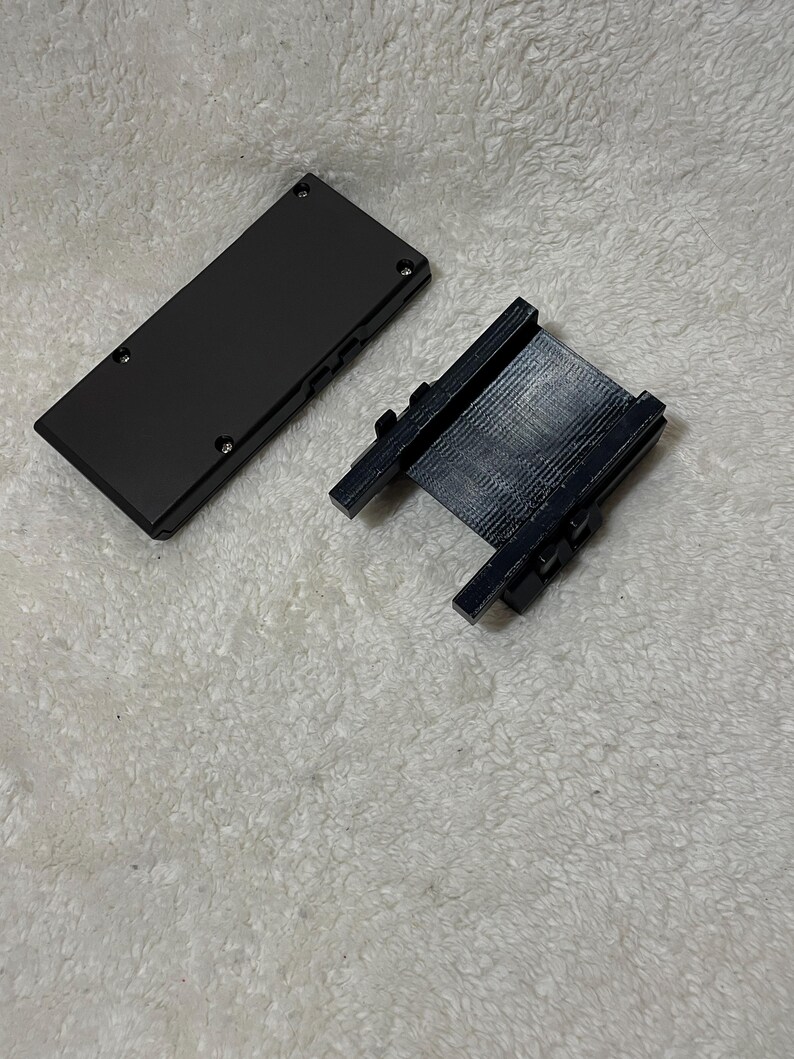 May include: Two black rectangular components, possibly electronic device parts, are displayed against a white, textured background. One component is a solid rectangle with screws, and the other is a frame-like structure.