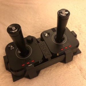 May include: A black Atari joystick with two controllers, each with a black joystick and a button labeled 'A' and 'B'. The joystick base has the word 'ATARI' printed on it in white letters.