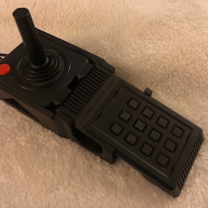 May include: A black video game controller featuring a vertical joystick, a single red circular button, and an integrated keypad labeled Video Touch Pad with twelve square buttons. A black cord extends from the rear of the device.