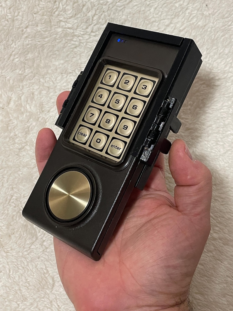 May include: A black and gold keypad device with a numeric keypad, a circular gold button, and the words "clear" and "enter". The device is held in a hand.