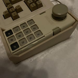 May include: A vintage beige computer keyboard with a numeric keypad and a joystick. The keyboard has beige keys and a beige frame. The numeric keypad has black numbers and symbols. The joystick is gray.