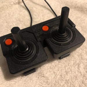 May include: A black two-player joystick controller with two orange buttons and two black joysticks. The controller is on a white fuzzy surface.