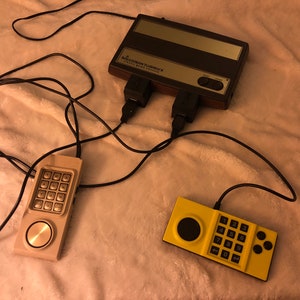 May include: A vintage Intellivision video game console with two controllers. The console is brown with a black screen and has the text "Intellivision" and "Video Game Console" printed on it. The controllers are beige and yellow with a keypad and a joystick.