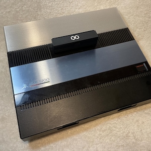May include: An Atari 5200 video game console. The console has a silver top with a black and blue center section. A black controller is on top, displaying the number "00". The Atari logo is visible.