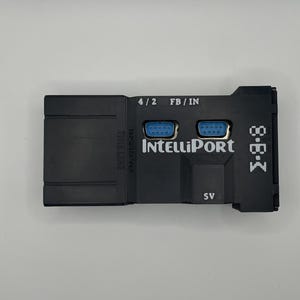 Op de afbeelding: Een zwart elektronisch apparaat met de tekst "IntelliPort" en "M-B-K" erop gedrukt. Het heeft twee blauwe poorten en de tekst "4/2 FB/IN" erboven. Het apparaat heeft ook de tekst "5V" erop gedrukt.