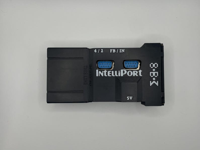 Op de afbeelding: Een zwart elektronisch apparaat met de tekst "IntelliPort" en "M-B-K" erop gedrukt. Het heeft twee blauwe poorten en de tekst "4/2 FB/IN" erboven. Het apparaat heeft ook de tekst "5V" erop gedrukt.