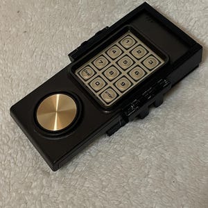 May include: A black electronic keypad with a gold-colored dial and a grid of beige buttons. The keypad has numbers 1-9, a clear button, and an enter button. The device is rectangular with a textured surface.