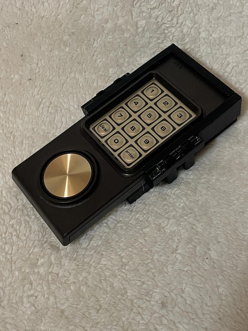 May include: A black electronic keypad with a gold-colored dial and a grid of beige buttons. The keypad has numbers 1-9, a clear button, and an enter button. The device is rectangular with a textured surface.