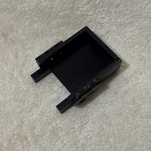 May include: A black rectangular box with a hinged lid and two small rectangular extensions on the sides. The box is open, revealing a dark interior. The object is set against a white, textured background.