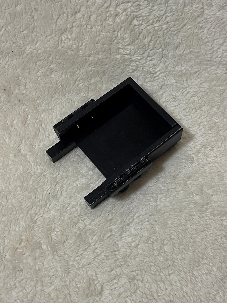 May include: A black rectangular box with a hinged lid and two small rectangular extensions on the sides. The box is open, revealing a dark interior. The object is set against a white, textured background.