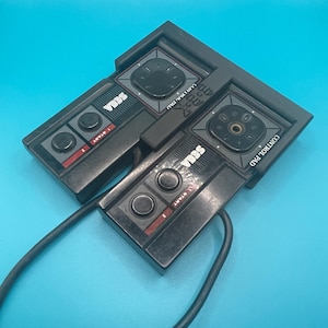 May include: Two black Sega Genesis controllers with cords attached. The controllers have a black and red color scheme and feature a directional pad, buttons, and a start button. The text "SEGA" is printed on the controllers.