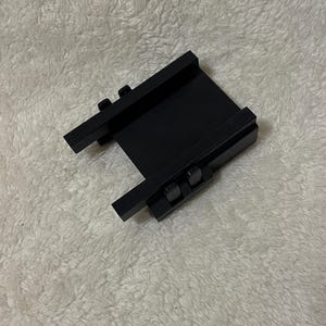 May include: A black metal bracket with two parallel rails and two smaller perpendicular rails. The bracket has two small, square-shaped fasteners on each of the perpendicular rails. The object is on a white, textured surface.