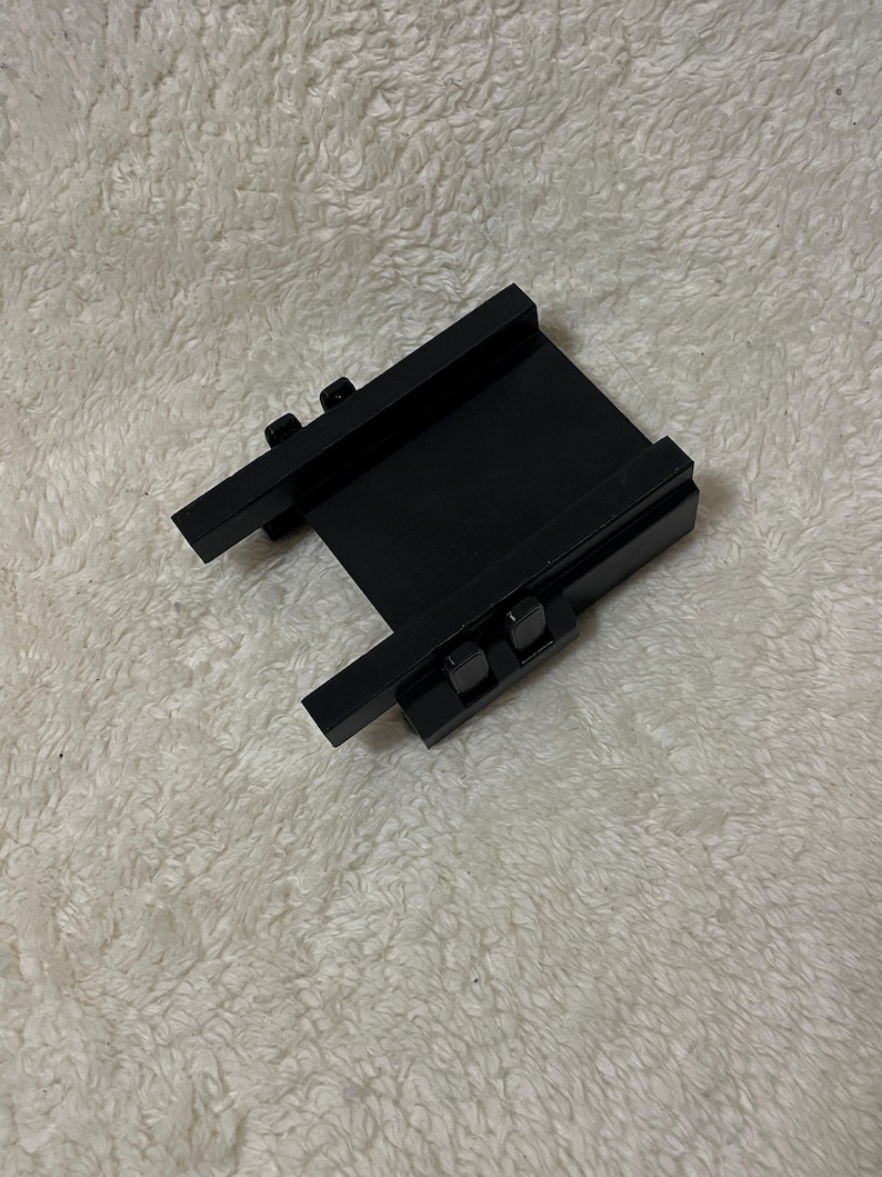 May include: A black metal bracket with two parallel rails and two smaller perpendicular rails. The bracket has two small, square-shaped fasteners on each of the perpendicular rails. The object is on a white, textured surface.