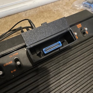 May include: Close-up of a black Atari 2600 video game console with a blue connector, power switch, and TV type selector. The console has a textured surface and the text "GAME RESET" is visible on the top right corner.